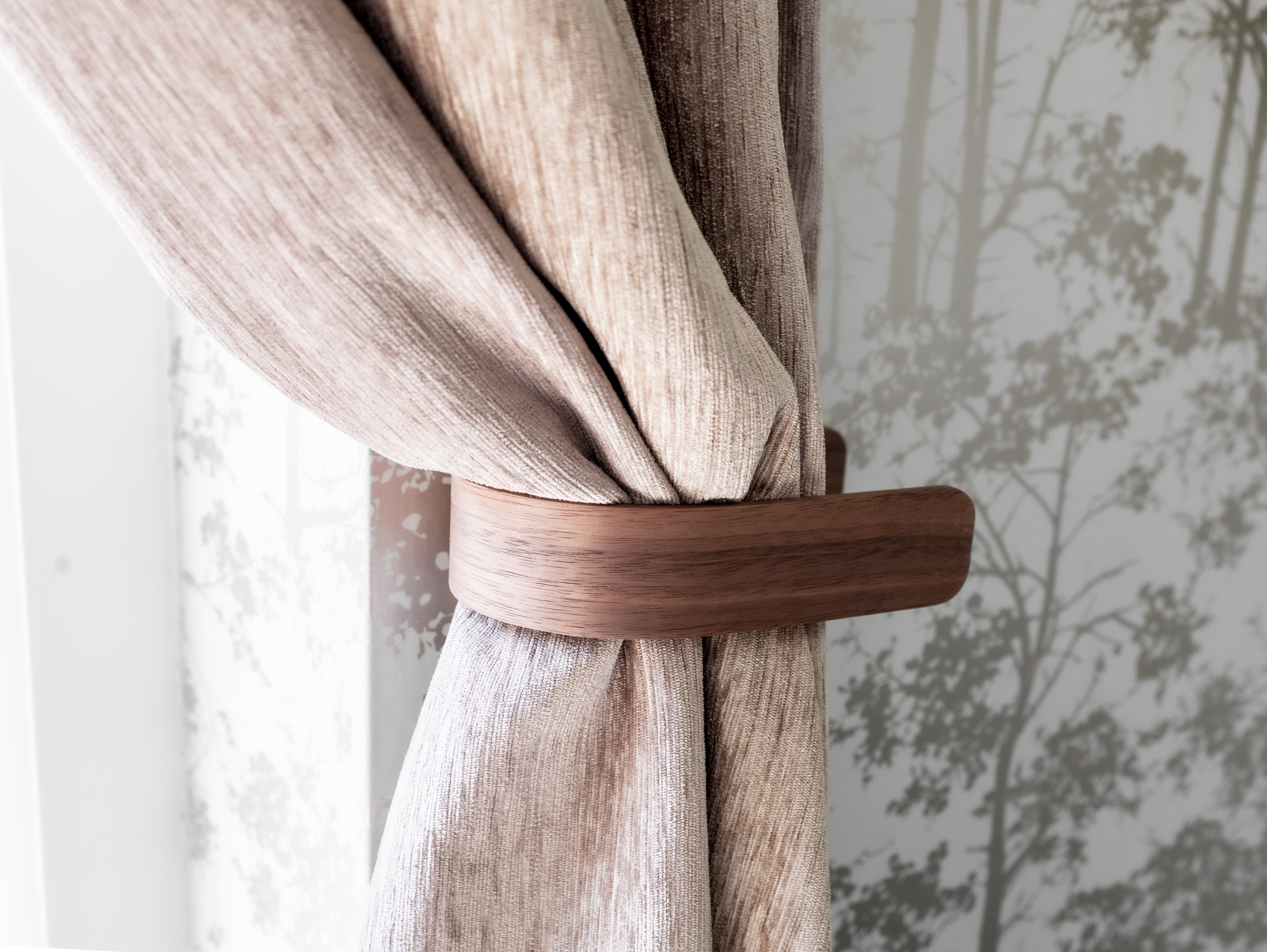 A walnut curtain holdback in brown color, attached to a curtain with a wall in the background featuring a subtle pattern.